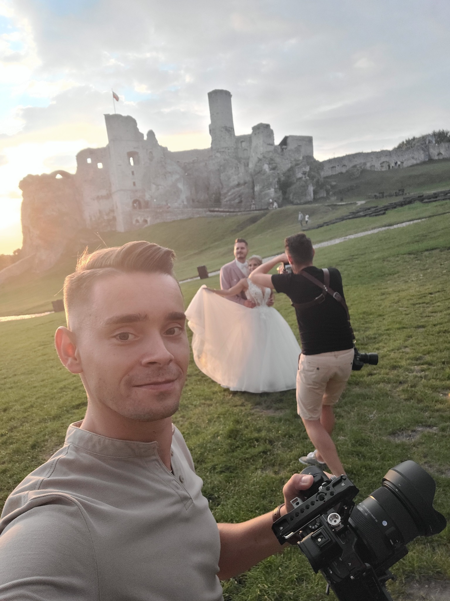Selfie kamerzysty z profesjonalnym sprzętem na tle pary młodej pozującej do zdjęcia ślubnego i średniowiecznego zamku w tle, podczas zachodu słońca.