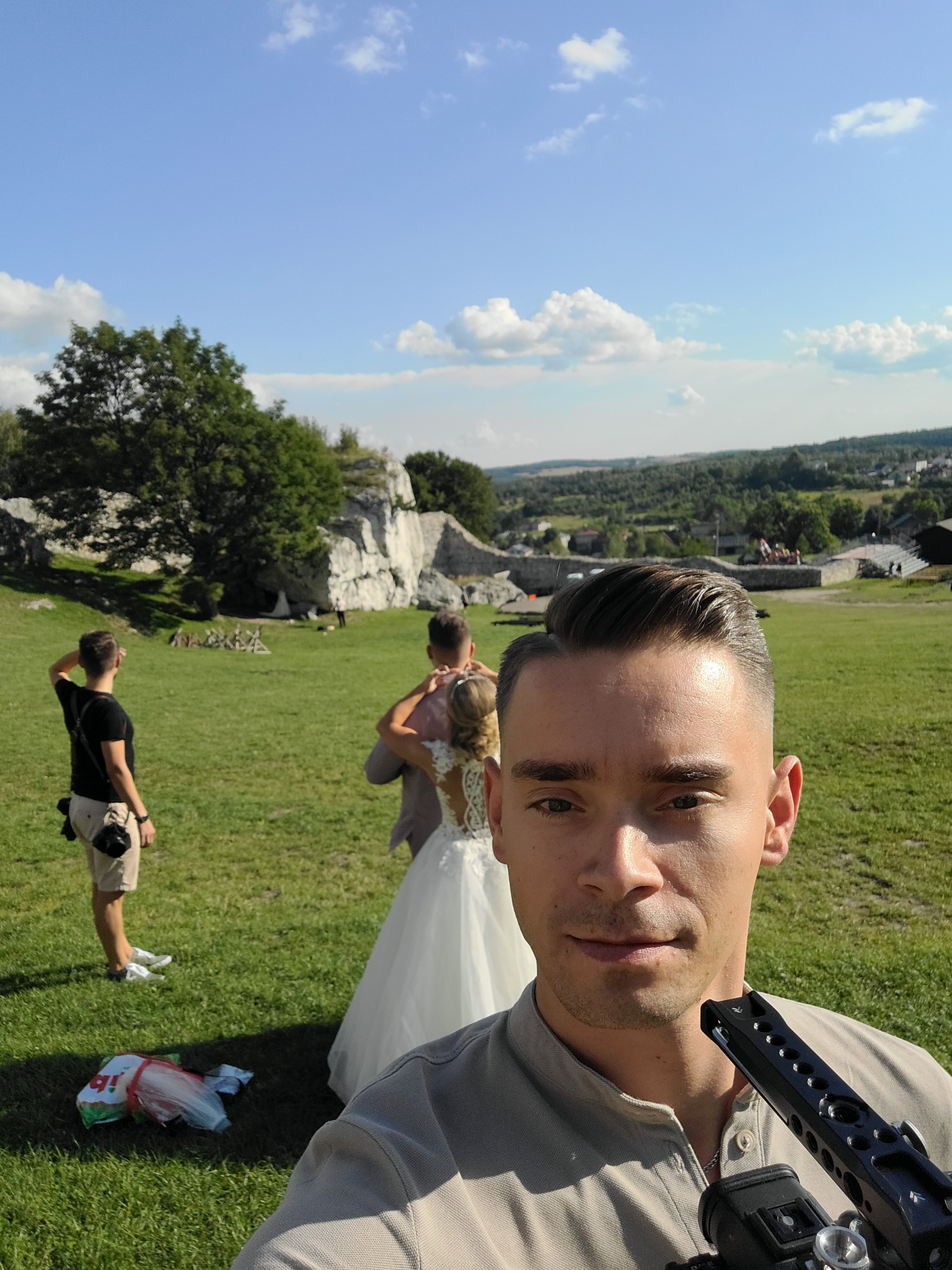 Selfie kamerzysty z profesjonalnym sprzętem na tle plenerowej sesji ślubnej w malowniczej scenerii z ruinami zamku, gdzie w oddali widać parę młodą i asystenta.