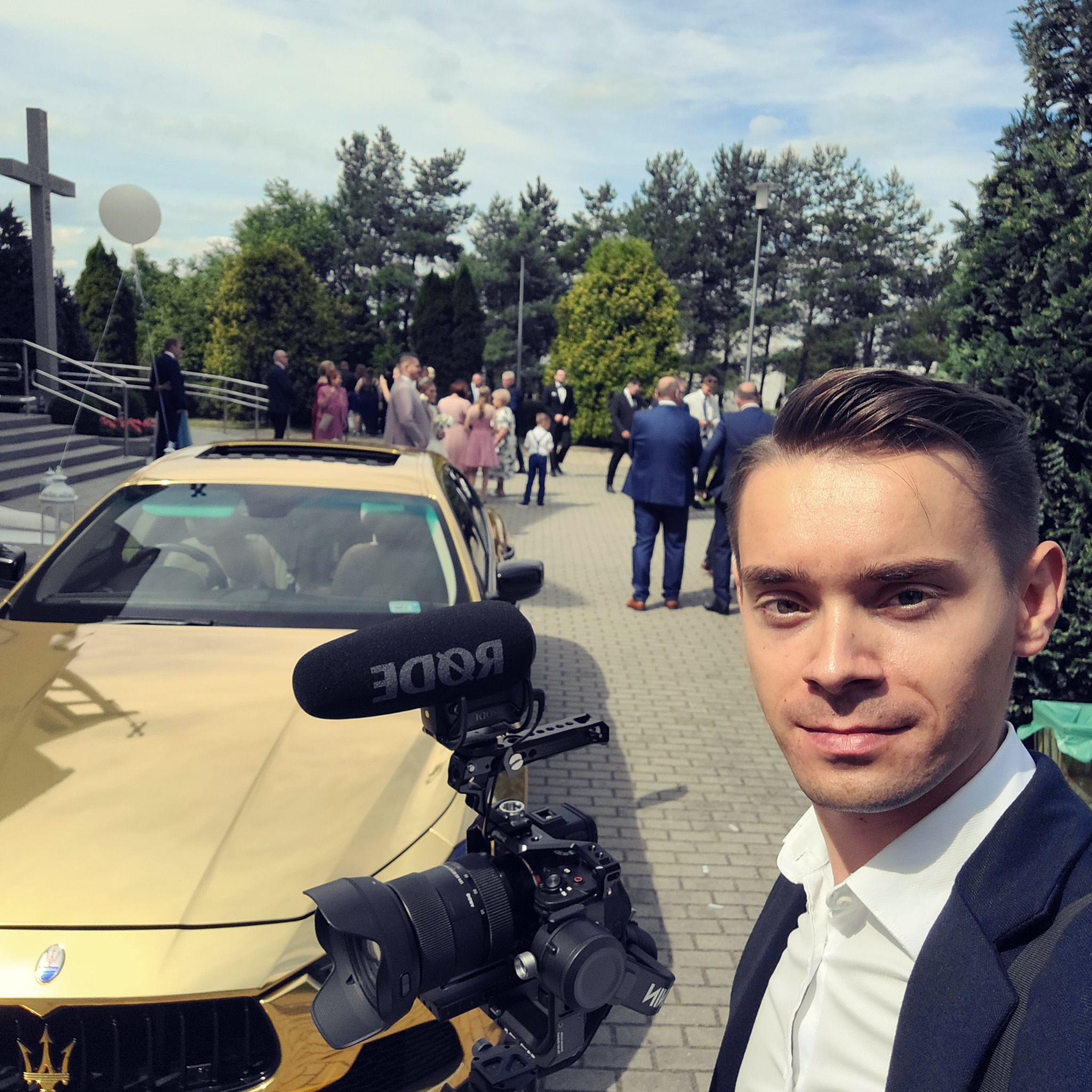 Selfie kamerzysty z profesjonalnym sprzętem (mikrofon Rode, kamera) na tle złotego samochodu i uroczystości ślubnej przed kościołem.