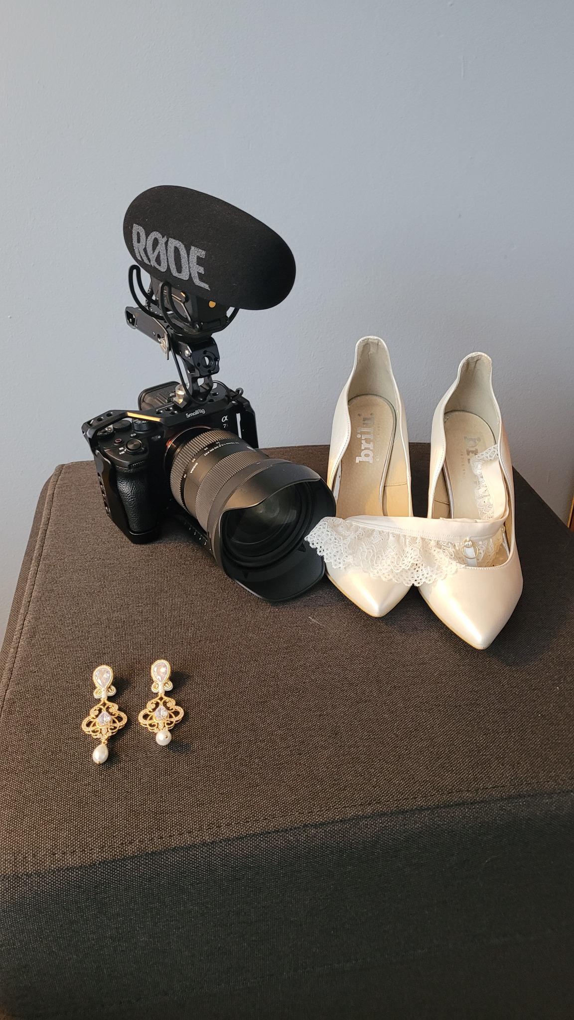 Profesjonalny sprzęt wideo z mikrofonem Rode, para eleganckich, kremowych szpilek z koronkową podwiązką oraz złote kolczyki z perłami, przygotowane do nagrania lub sesji zdjęciowej ślubu.