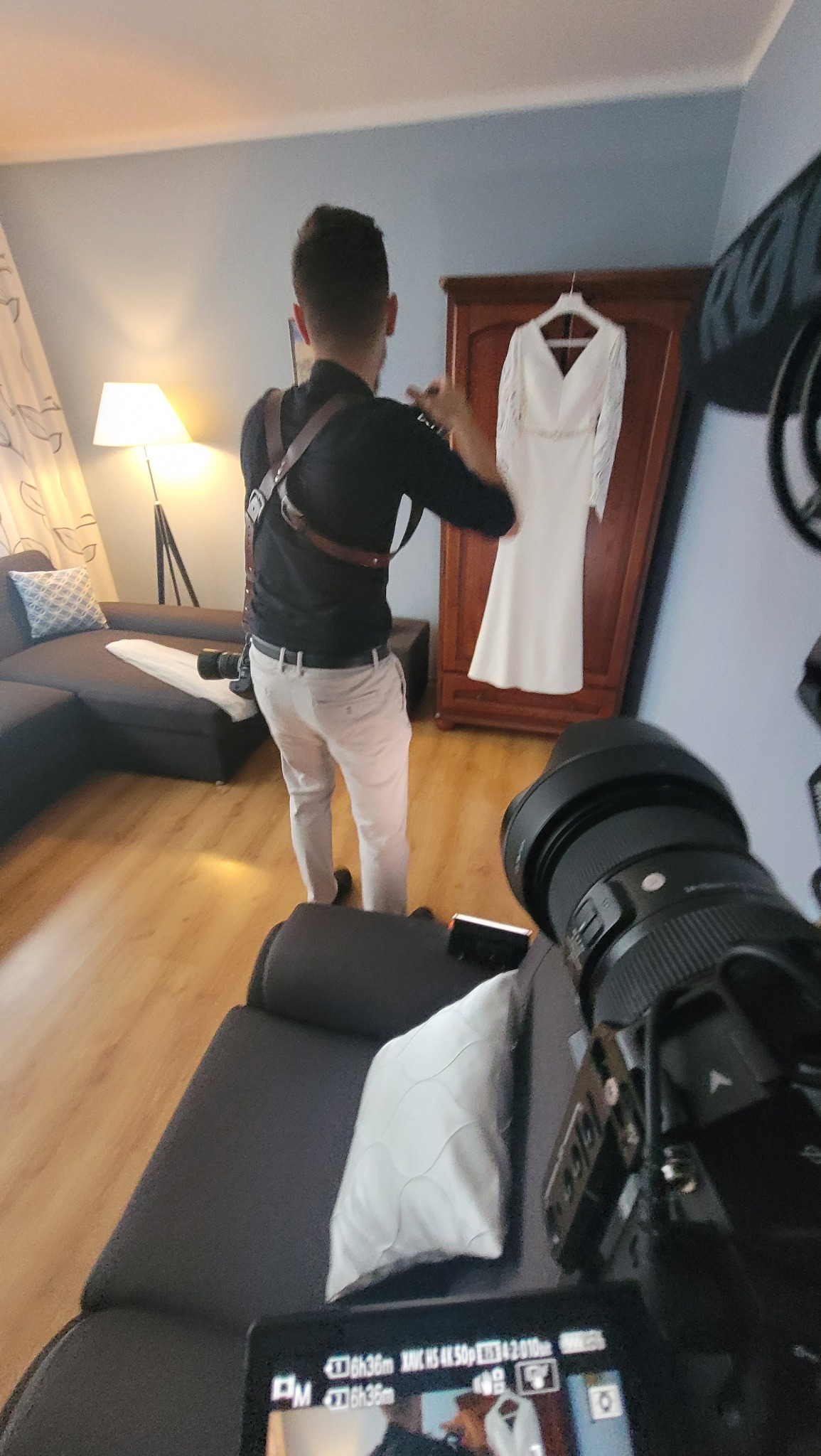 Kamerzysta w trakcie pracy, filmuje białą suknię ślubną wiszącą na szafie w pokoju, widoczny ekran kamery z podglądem kadru.