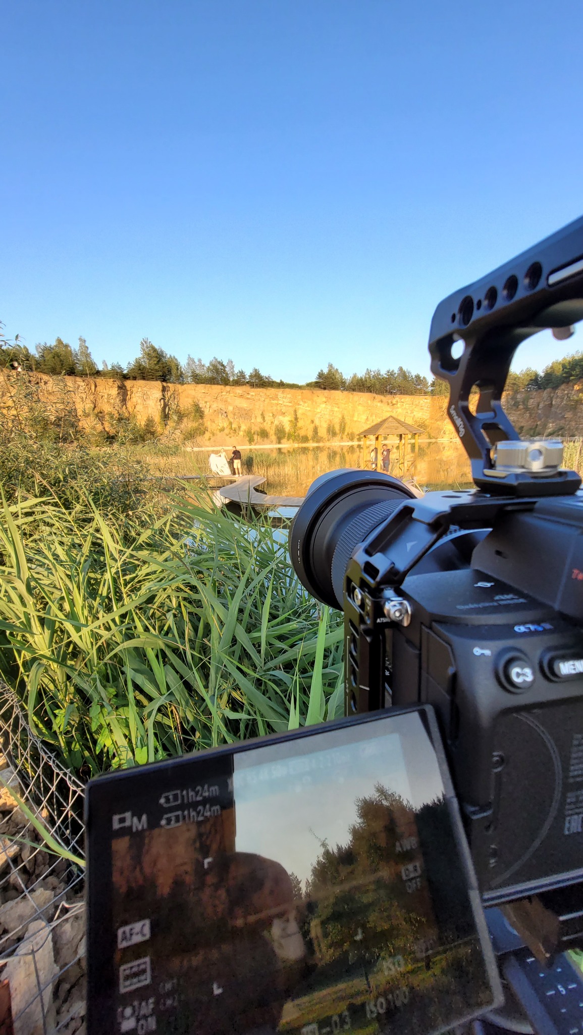 Kamera z osprzętem ustawiona do nagrywania plenerowej sesji w Dąbrowie Górniczej; w tle widoczna para na pomoście nad jeziorem i altana.