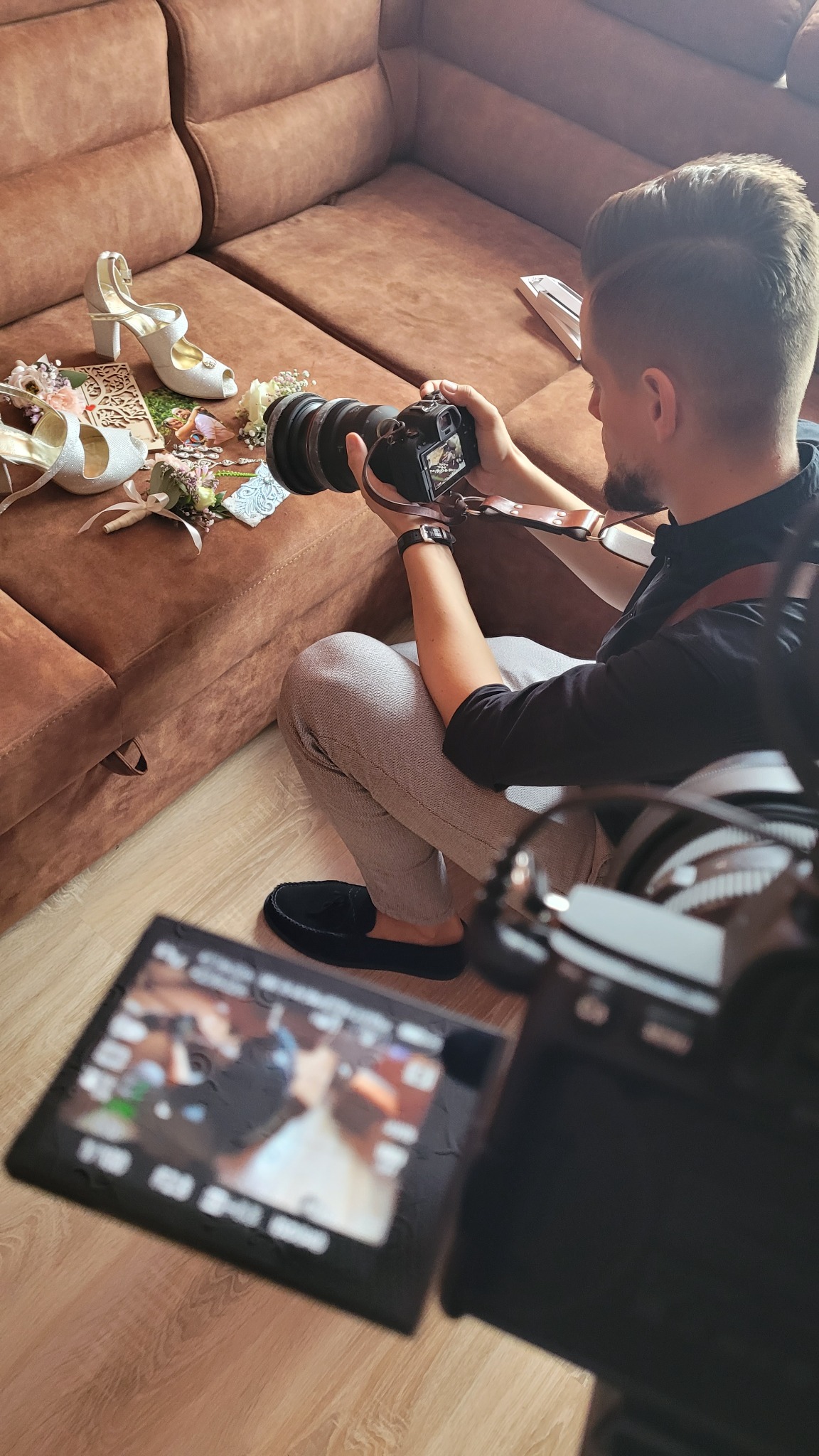 Kamerzysta fotografuje buty ślubne, bukiecik i ozdobne pudełko na brązowej kanapie, widok zza drugiego aparatu z podglądem kadru
