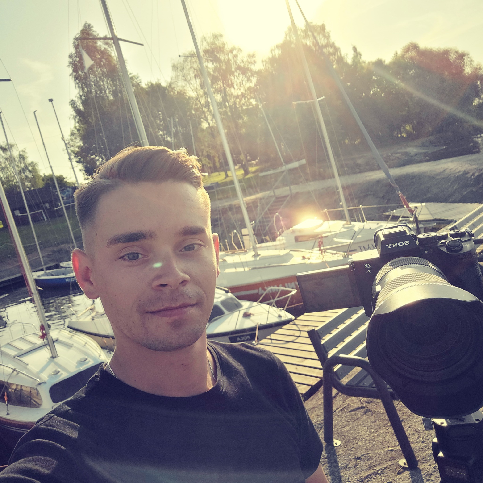 Autoportret mężczyzny z profesjonalnym aparatem Sony Alpha na tle przystani jachtowej w słoneczny dzień.