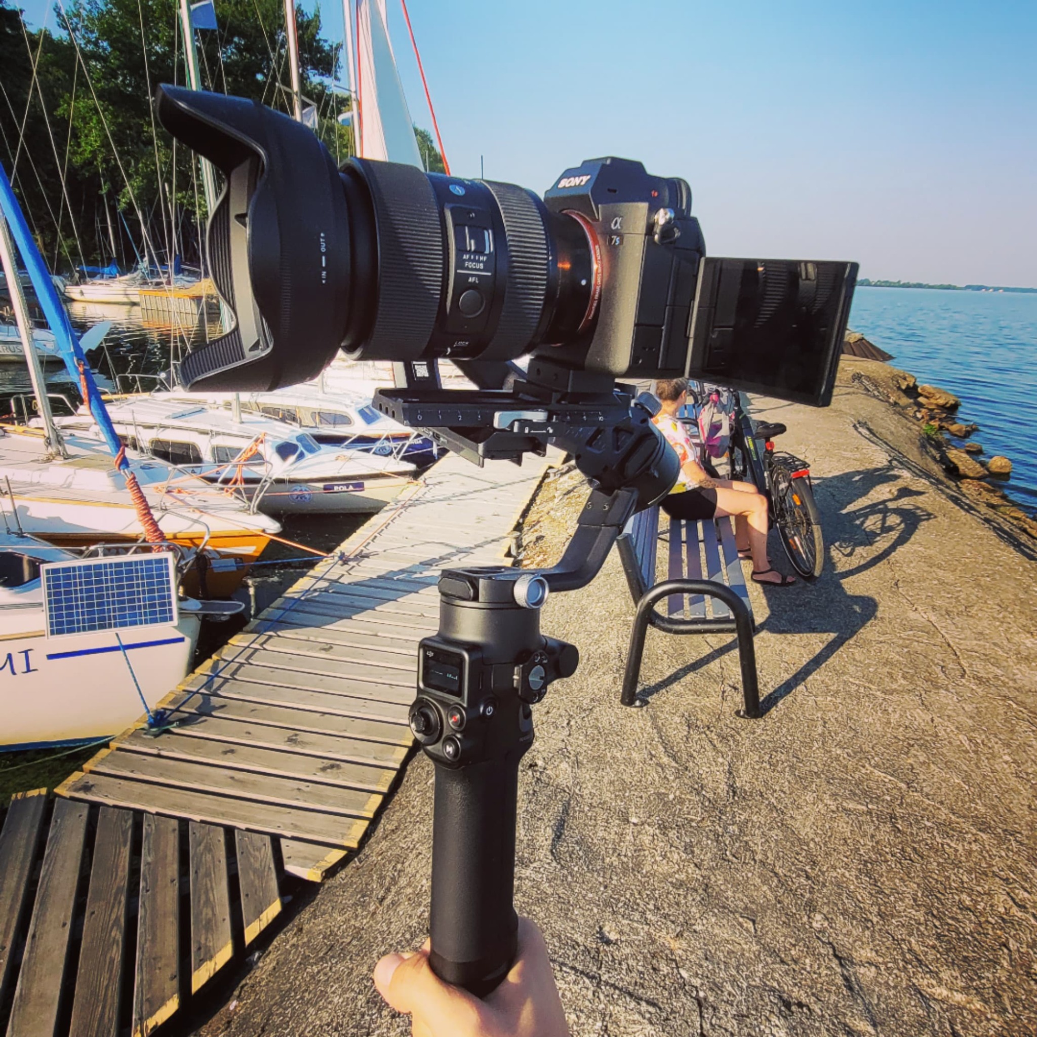 Kamera Sony Alpha 7S zamontowana na gimbalu, uchwycona z perspektywy pierwszej osoby na tle przystani z jachtami i osoby siedzącej na ławce nad wodą.