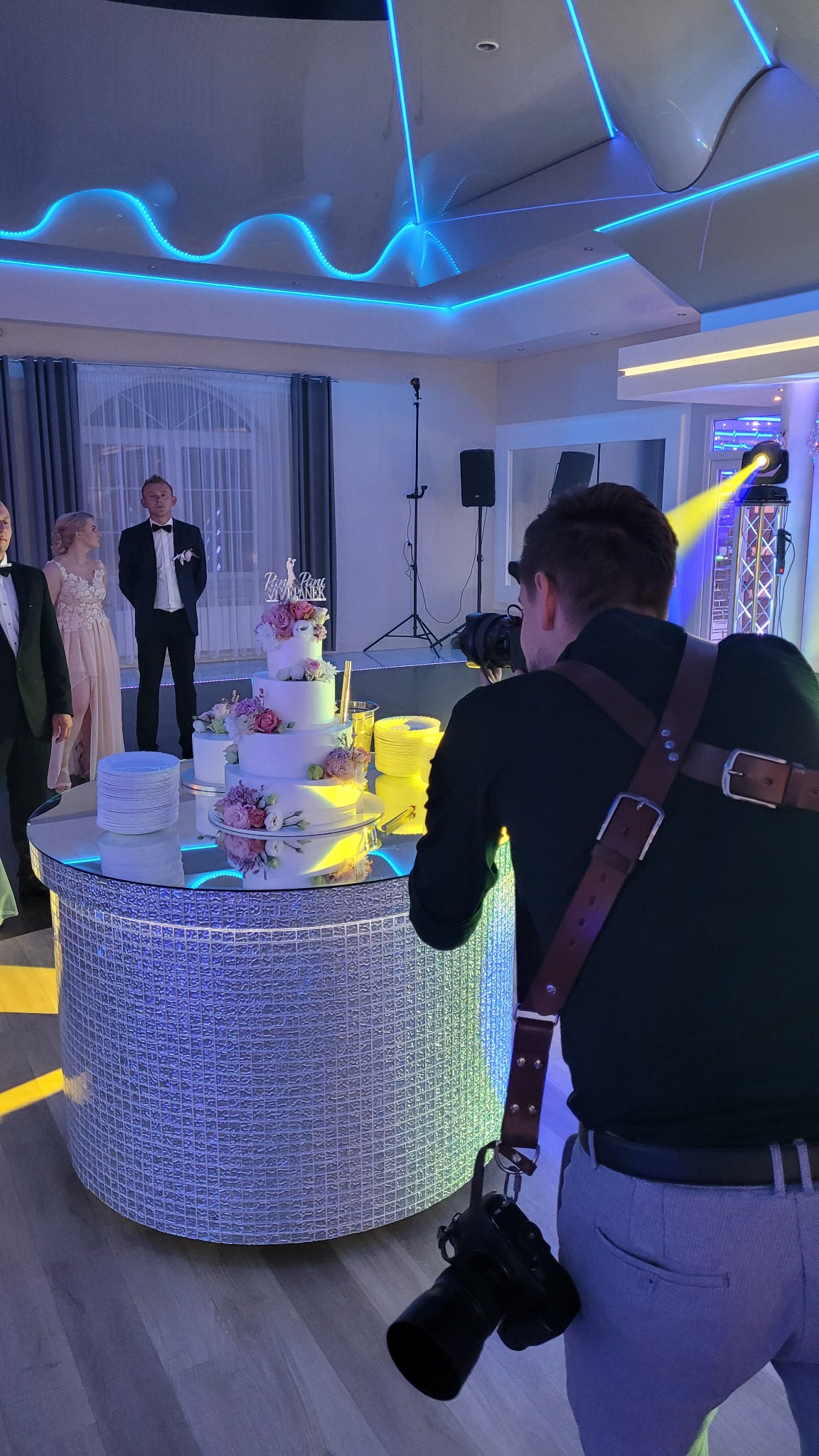 Fotograf w trakcie pracy, filmujący parę młodą i tort weselny w oświetlonym sali bankietowej z dekoracjami.