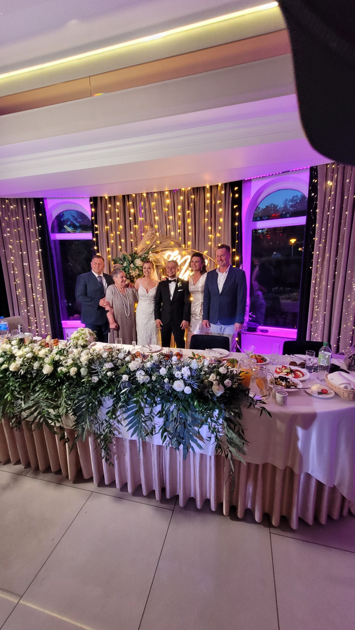 Zdjęcie nowożeńców z gośćmi weselnymi za stołem udekorowanym kwiatami i jedzeniem, oświetlonym girlandami żarówek i fioletowym światłem
