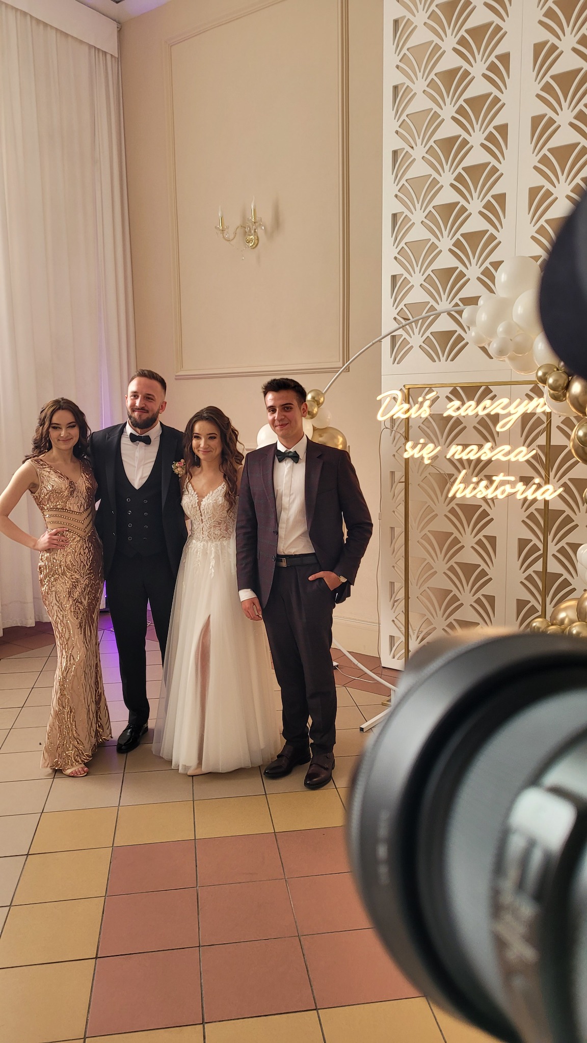 Grupa elegancko ubranych osób, w tym panna młoda w sukni z rozcięciem, pozuje na tle dekoracji ślubnej z balonami i napisem neonowym 'Dziś zaczyna się nasza historia', widoczny obiektyw aparatu.