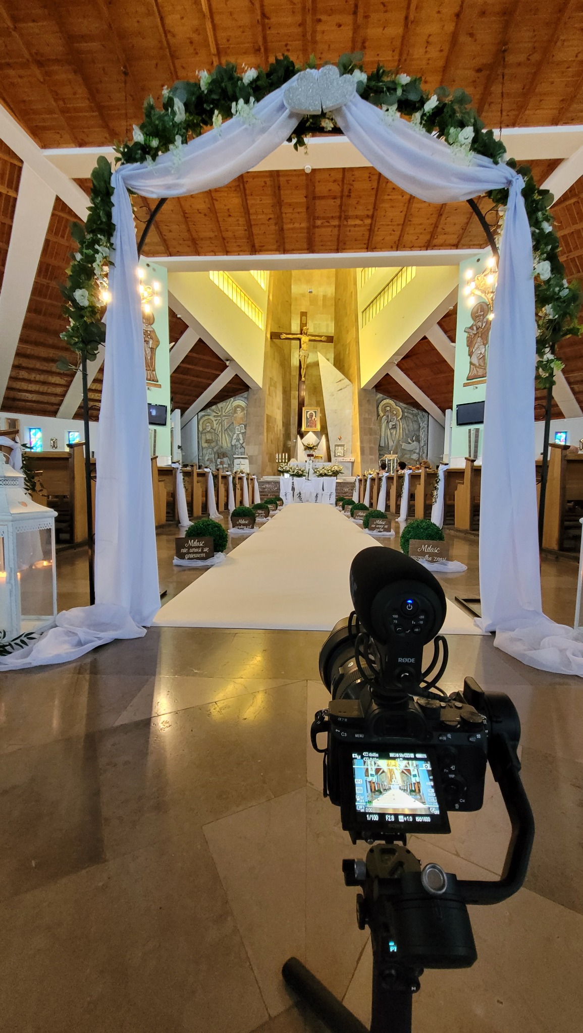 Kamera z mikrofonem na stabilizatorze ustawiona w kościele przed ślubnym kobiercem i dekoracjami kwiatowymi na łuku, widok z perspektywy kamerzysty.