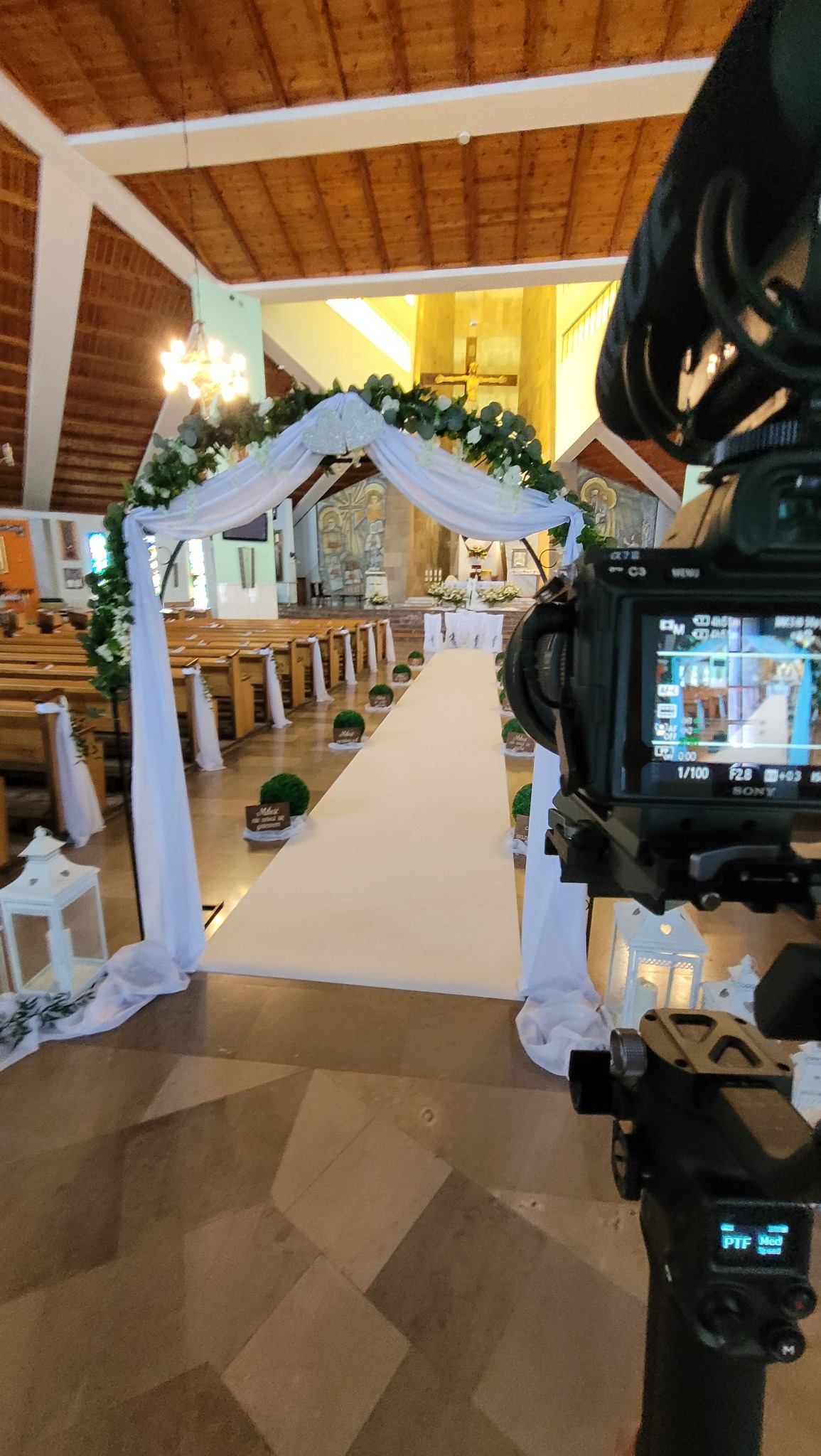 Kamera Sony ustawiona w kościele, nagrywająca przygotowania do ceremonii ślubnej, widoczny biały dywan, dekoracje kwiatowe i ławki.