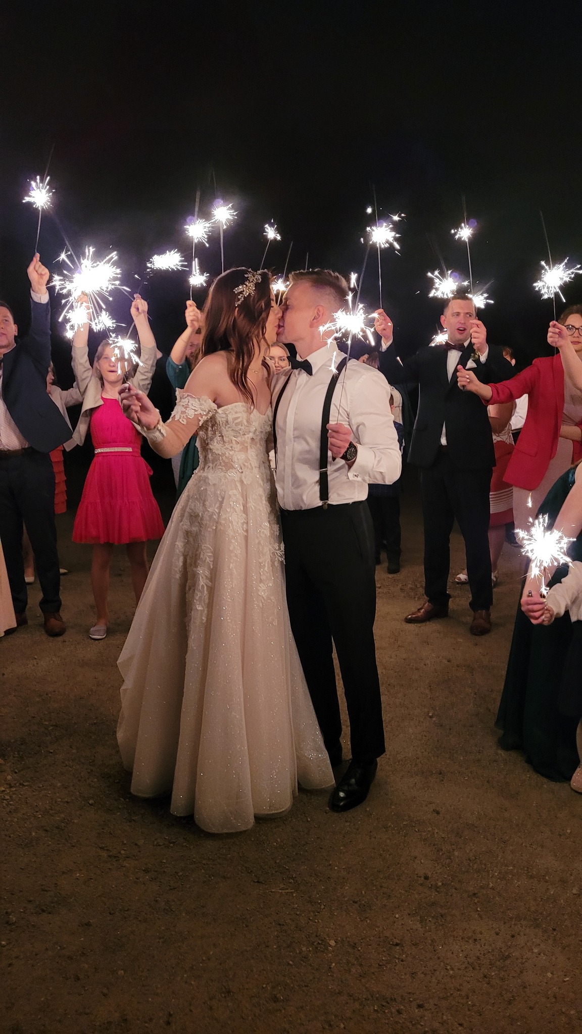 Pocałunek nowożeńców otoczonych gośćmi z zimnymi ogniami, suknia ślubna z koronkowymi zdobieniami i odkrytymi ramionami, mężczyzna w białej koszuli z czarnymi szelkami i muszką.