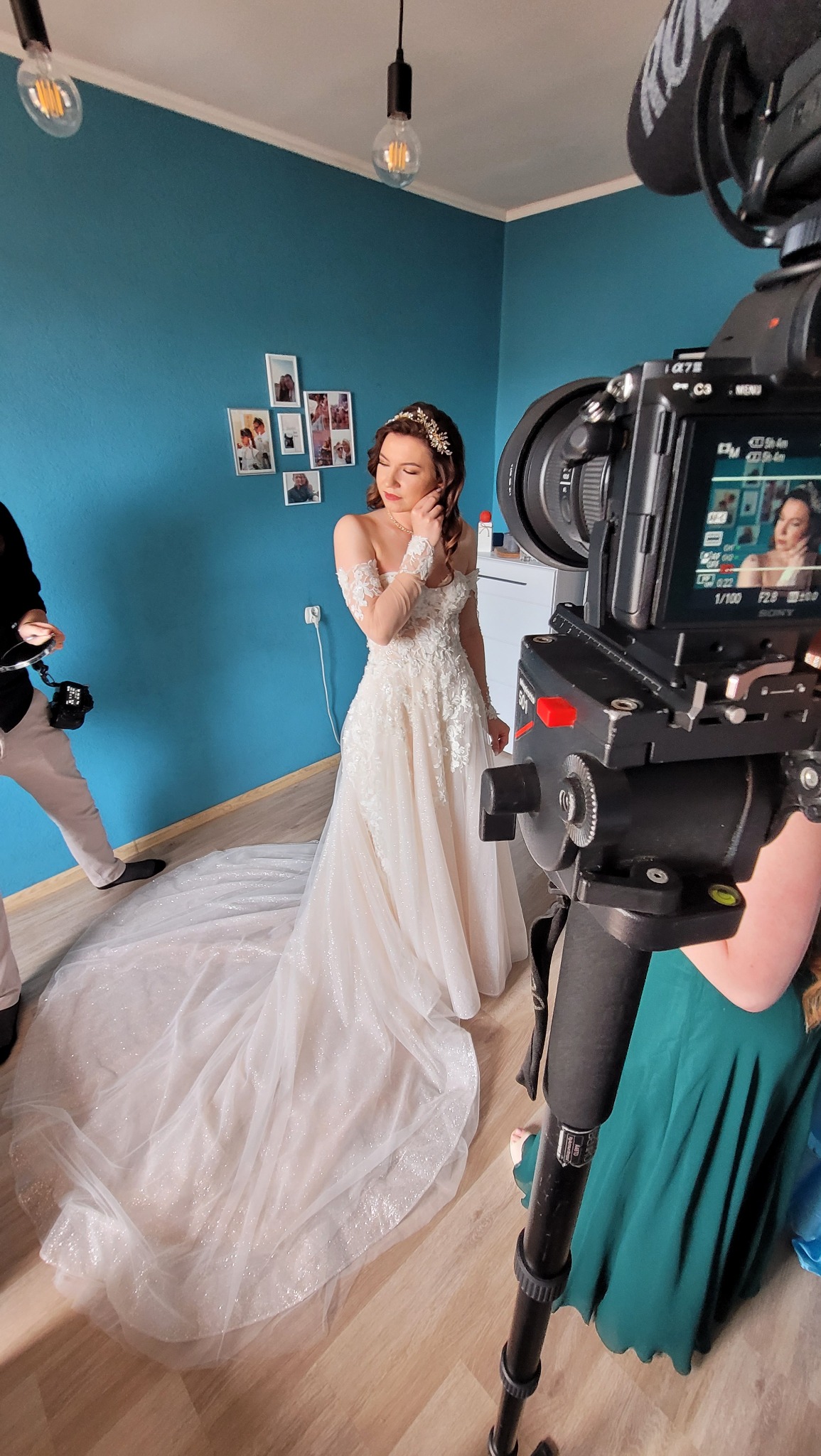 Panna młoda w koronie i sukni ślubnej pozuje przed kamerą filmową z mikrofonem, w tle błękitna ściana z ramkami zdjęć.