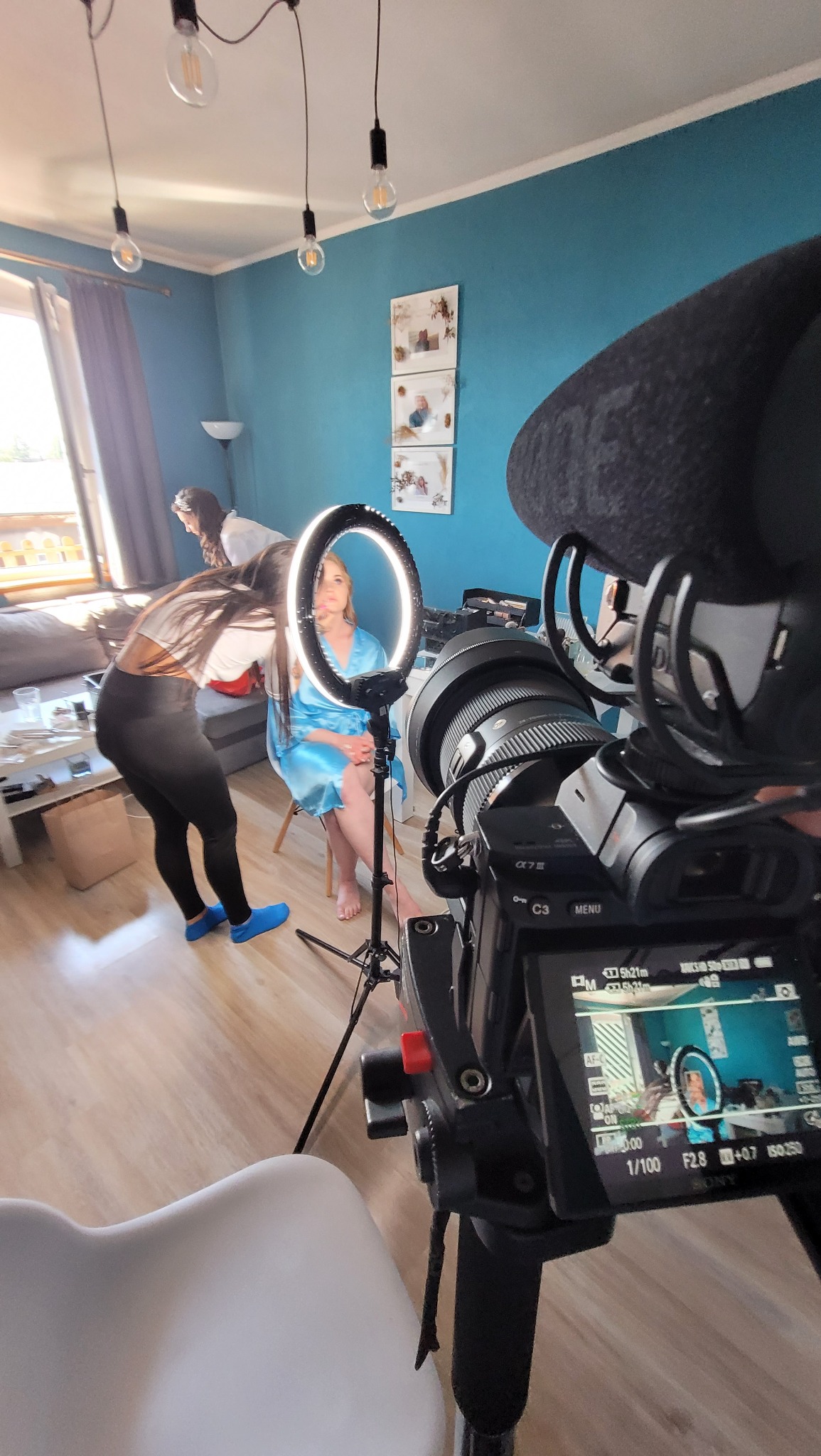 Kamera z mikrofonem na statywie rejestruje scenę przygotowań modelki do sesji zdjęciowej, oświetloną lampą pierścieniową; w tle wizażystka poprawia makijaż.