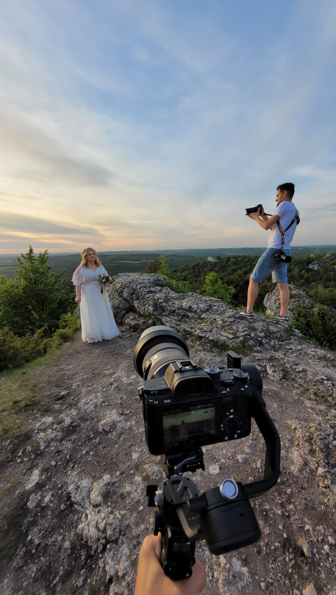 Ujęcie zza kamery na stabilizatorze, ukazujące fotografa robiącego zdjęcia pannie młodej na tle malowniczego krajobrazu o zachodzie słońca.