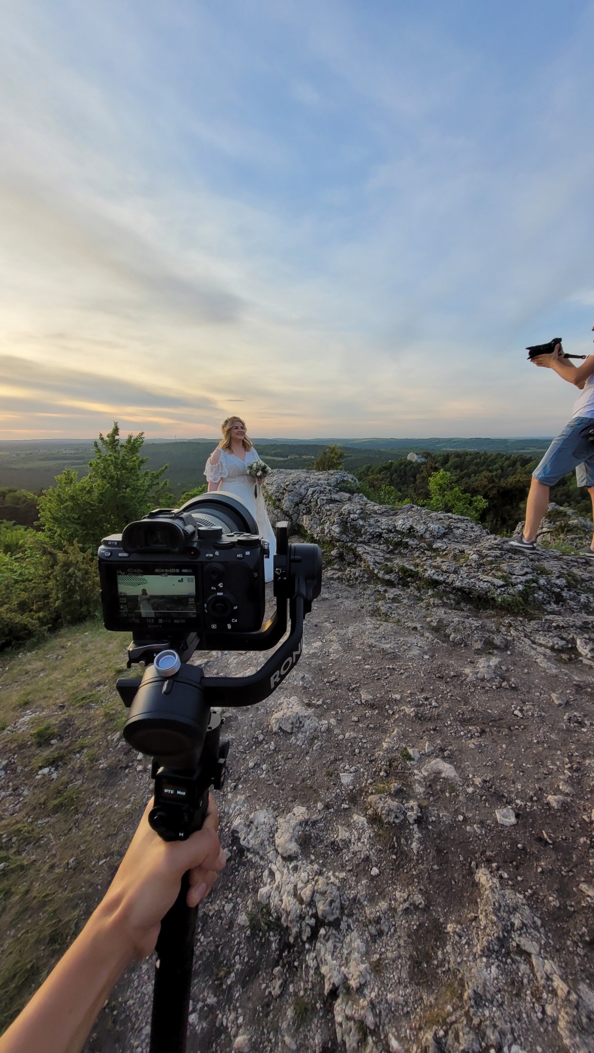 Kamera z gimbalem nagrywa uśmiechniętą pannę młodą w białej sukni, stojącą na skalistym wzgórzu z widokiem na zielone lasy i zachodzące słońce; drugi kamerzysta w tle.