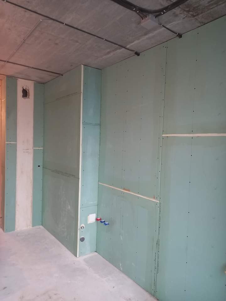 Wnętrze w trakcie prac wykończeniowych: ściany obłożone zielonymi płytami kartonowo-gipsowymi, widoczne instalacje wodne i elektryczne, surowy betonowy sufit z przewodami.