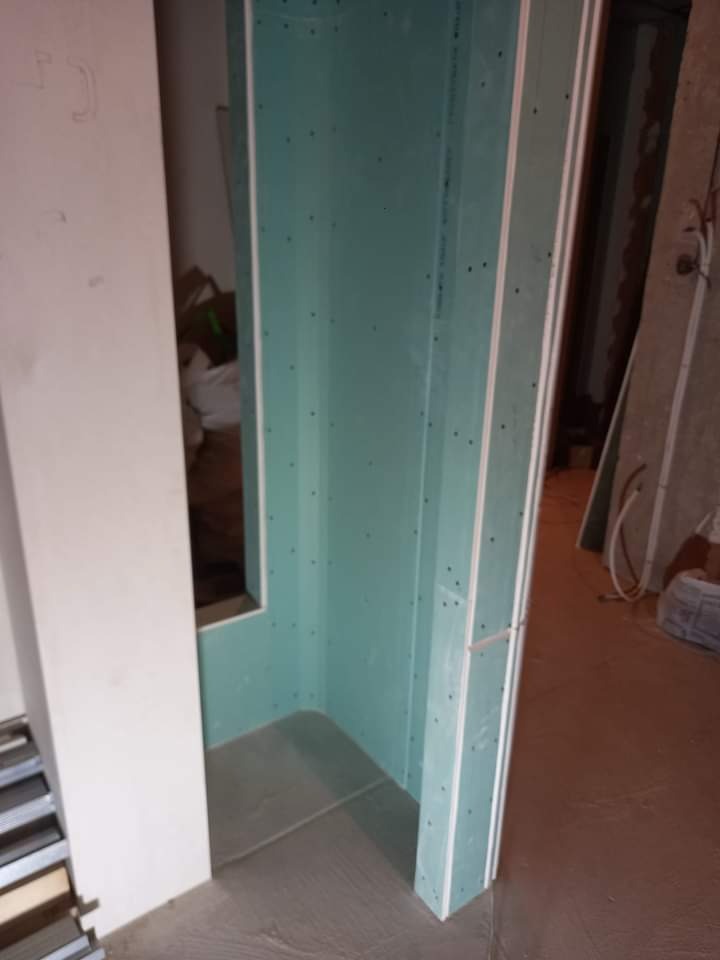 Montaż zielonej płyty gipsowo-kartonowej tworzącej narożnik w pomieszczeniu w trakcie prac wykończeniowych, widoczne profile i szpachlowana podłoga.