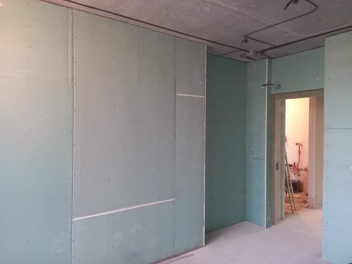 Wnętrze w trakcie montażu ścian z płyt kartonowo-gipsowych; widoczne łączenia płyt, surowy betonowy sufit z instalacją elektryczną, oraz fragment pomieszczenia z drabiną i instalacją hydrauliczną...