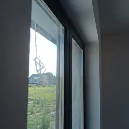 Widok przez okno z czarną ramą na dom z ogrodem; na szybie zwisająca taśma malarska, ściana w szarym kolorze.