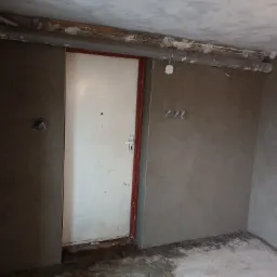 Wnętrze w trakcie prac wykończeniowych, ściany pokryte świeżo nałożoną szarą gładzią, widoczne rury pod sufitem i stara, biała metalowa futryna drzwiowa z czerwonym obramowaniem.