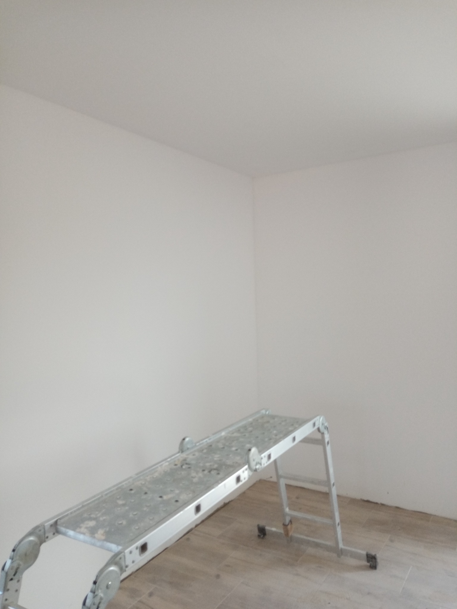 Pomalowane na biało wnętrze pokoju z aluminiowym podestem roboczym na kółkach, widoczny fragment podłogi imitującej drewno.