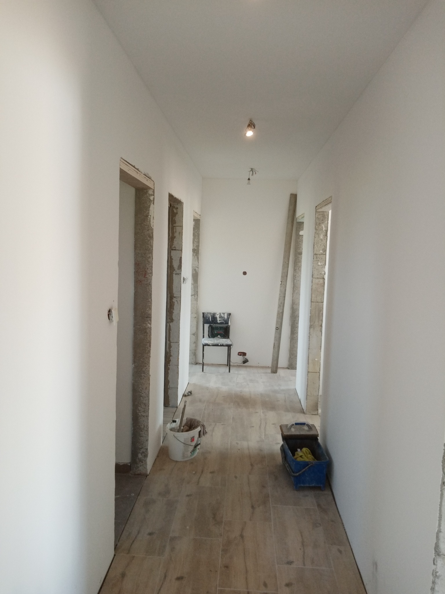 Korytarz w trakcie malowania na biało, widoczne narzędzia malarskie, surowe ościeżnice i podłoga imitująca drewno.