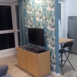 Telewizor zamontowany na ścianie z tapetą w liście monstery, stojący na szafce RTV z czarnym blatem w pokoju z drewnianą podłogą i zasłoną.