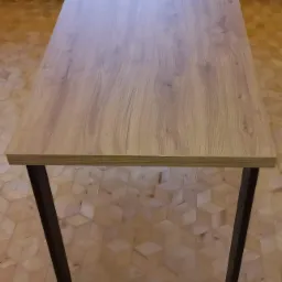 Stół z blatem imitującym jasne drewno i czarnymi, metalowymi nogami w kształcie trapezu, na tle wzorzystej podłogi.