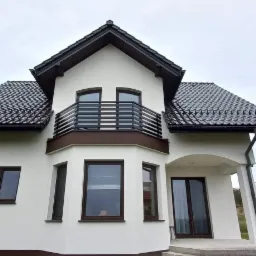 Nowoczesny dom jednorodzinny z białymi ścianami, brązowymi oknami i czarnym dachem; balkon z poziomymi, metalowymi barierkami.