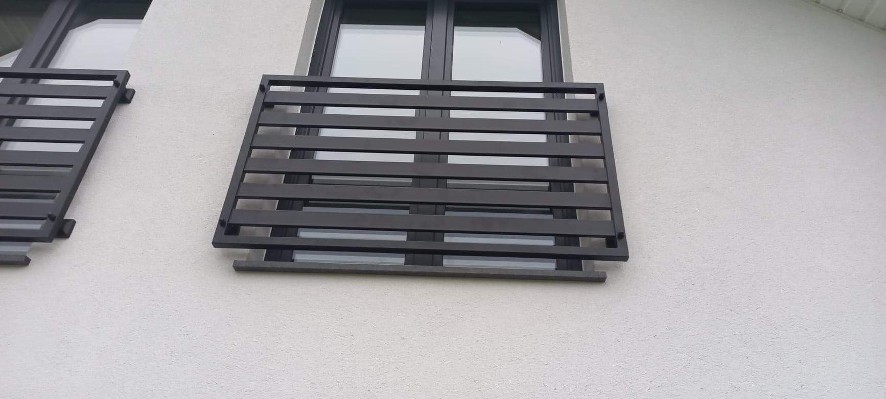 Zewnętrzna, metalowa balustrada okienna w kolorze grafitowym, z poziomymi szczeblami, zamontowana na elewacji budynku z widocznym fragmentem okna.