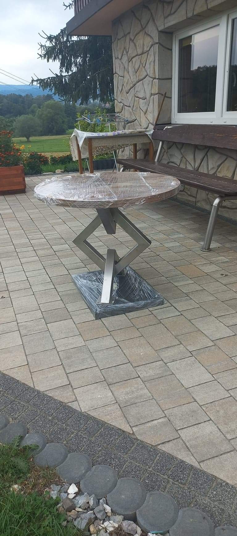 Okrągły stół z drewnianym blatem i geometryczną podstawą ze stali nierdzewnej, zabezpieczony folią ochronną, na tarasie z kostki brukowej.