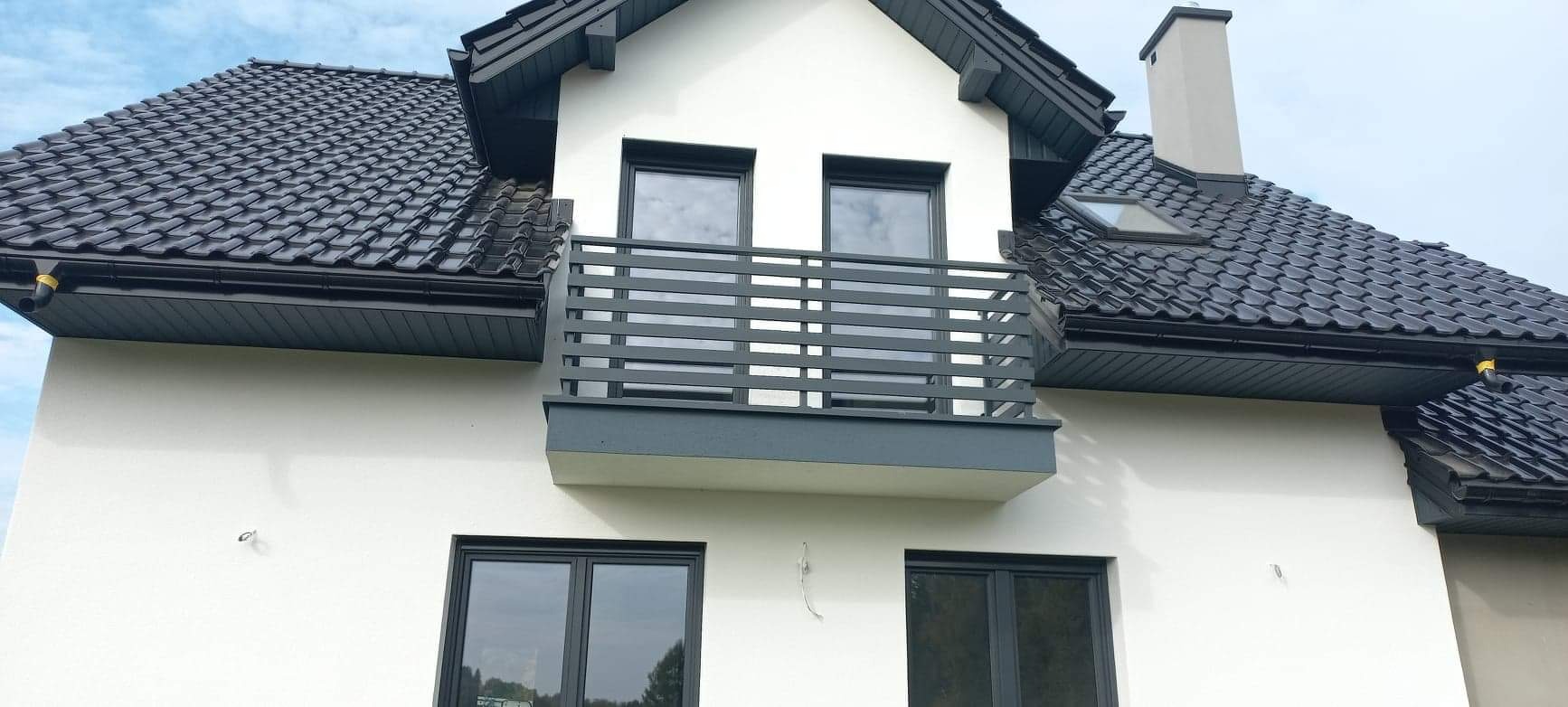 Nowoczesny dom z białymi ścianami i ciemnoszarą balustradą balkonową o poziomym układzie szczebli, pasująca do koloru dachu i okien.