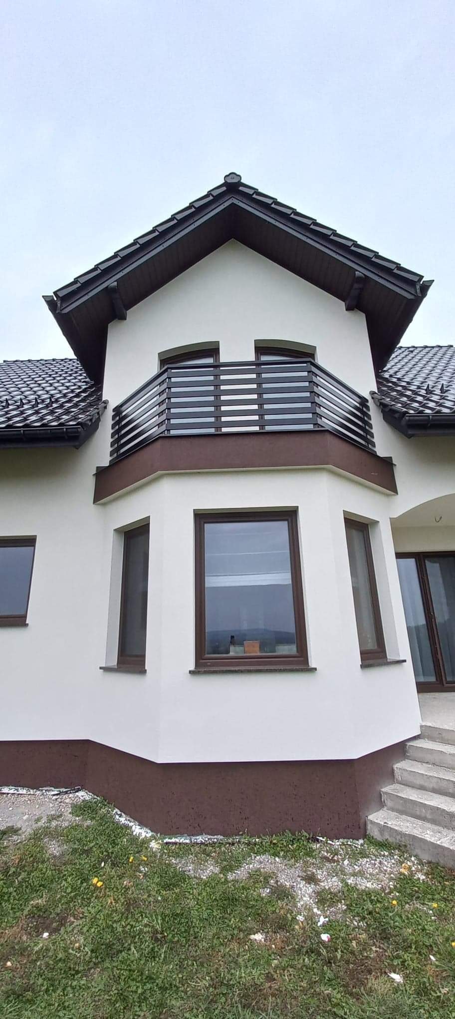 Nowoczesny dom z balkonem zabezpieczonym poziomą, metalową balustradą w kolorze ciemnego brązu, dopasowanym do stolarki okiennej i cokołu budynku.