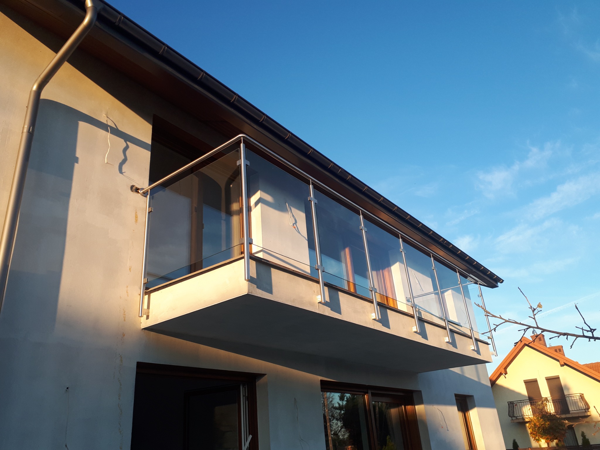 Nowoczesny balkon z balustradą szklaną i stalowymi elementami mocującymi, oświetlony popołudniowym słońcem, widok od dołu na tle błękitnego nieba i fragmentu sąsiedniego domu.