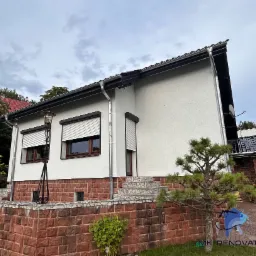 Biała elewacja domu z brązowymi oknami i roletami zewnętrznymi, widoczne rynny, kamienna podmurówka i ozdobne rośliny w ogrodzie, oświetlenie zewnętrzne w stylu retro.