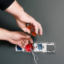 ArtLine - Elektryk przycina przewód obok gniazdka USB, widoczne trzy otwory na gniazdka w szarej ścianie, jeden z przewodami w puszce, używane szczypce z pomarańczowymi rączkami.