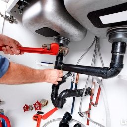 ArtLine - Hydraulik dokręca czerwoną żabką czarną rurę odpływową pod zlewem; widoczne liczne rury, zawory, wężyki i syfon; ciasna przestrzeń pod zlewem wypełniona instalacją hydrauliczną.