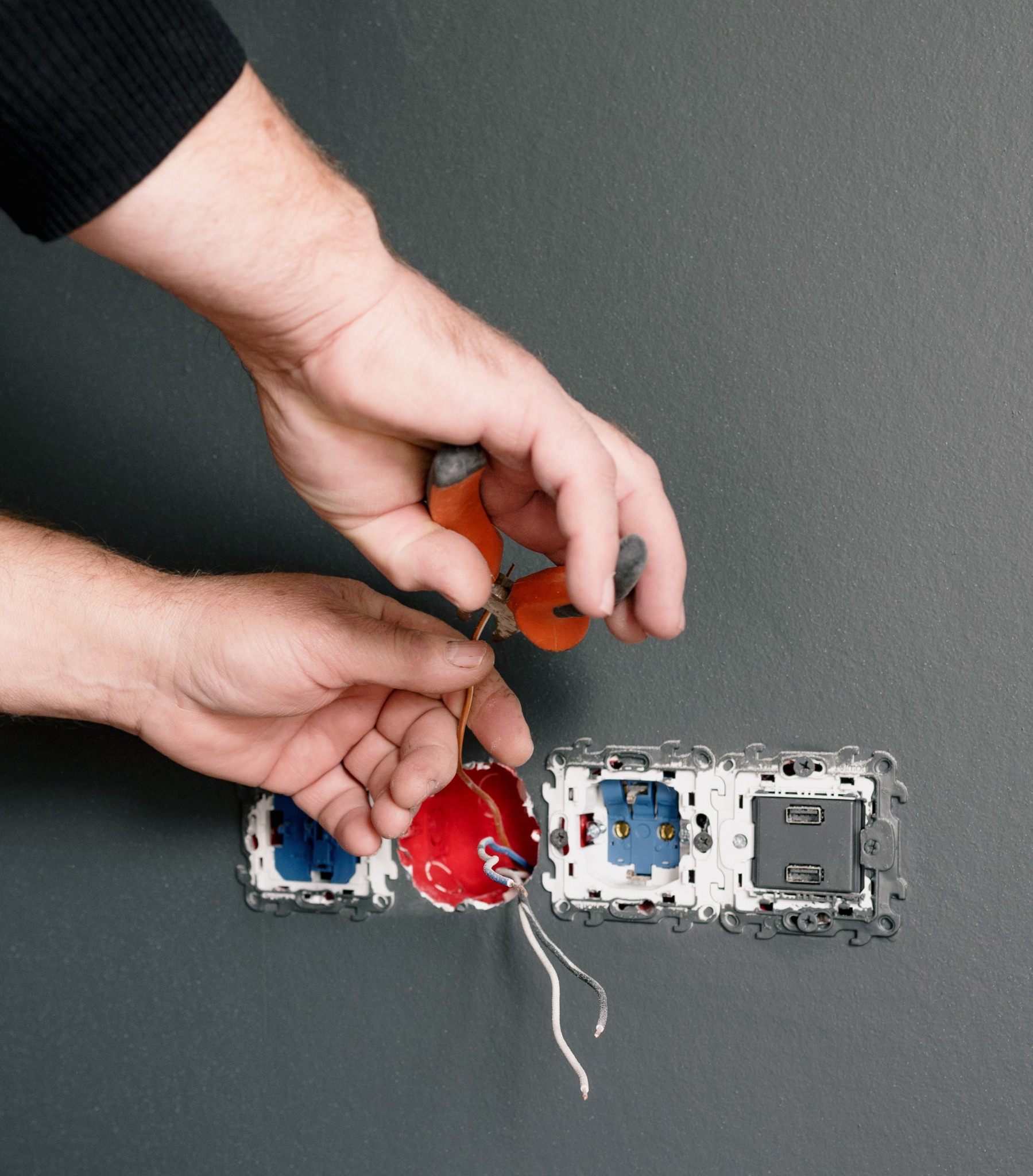 Elektryk przycina przewód obok gniazdka USB, widoczne trzy otwory na gniazdka w szarej ścianie, jeden z przewodami w puszce, używane szczypce z pomarańczowymi rączkami.