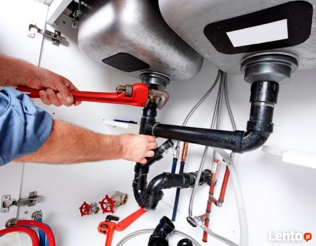 Hydraulik dokręca czerwoną żabką czarną rurę odpływową pod zlewem; widoczne liczne rury, zawory, wężyki i syfon; ciasna przestrzeń pod zlewem wypełniona instalacją hydrauliczną.