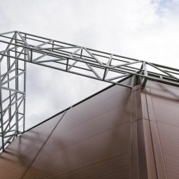 Yauheni Novak - Szkielet stalowej konstrukcji budynku przemysłowego z brązową elewacją panelową, widziany z dołu na tle pochmurnego nieba.