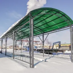Zadaszenie z zielonego poliwęglanu na metalowej konstrukcji, pokryte śniegiem, na tle zimowego krajobrazu z zaparkowanymi ciężarówkami i kominem przemysłowym.