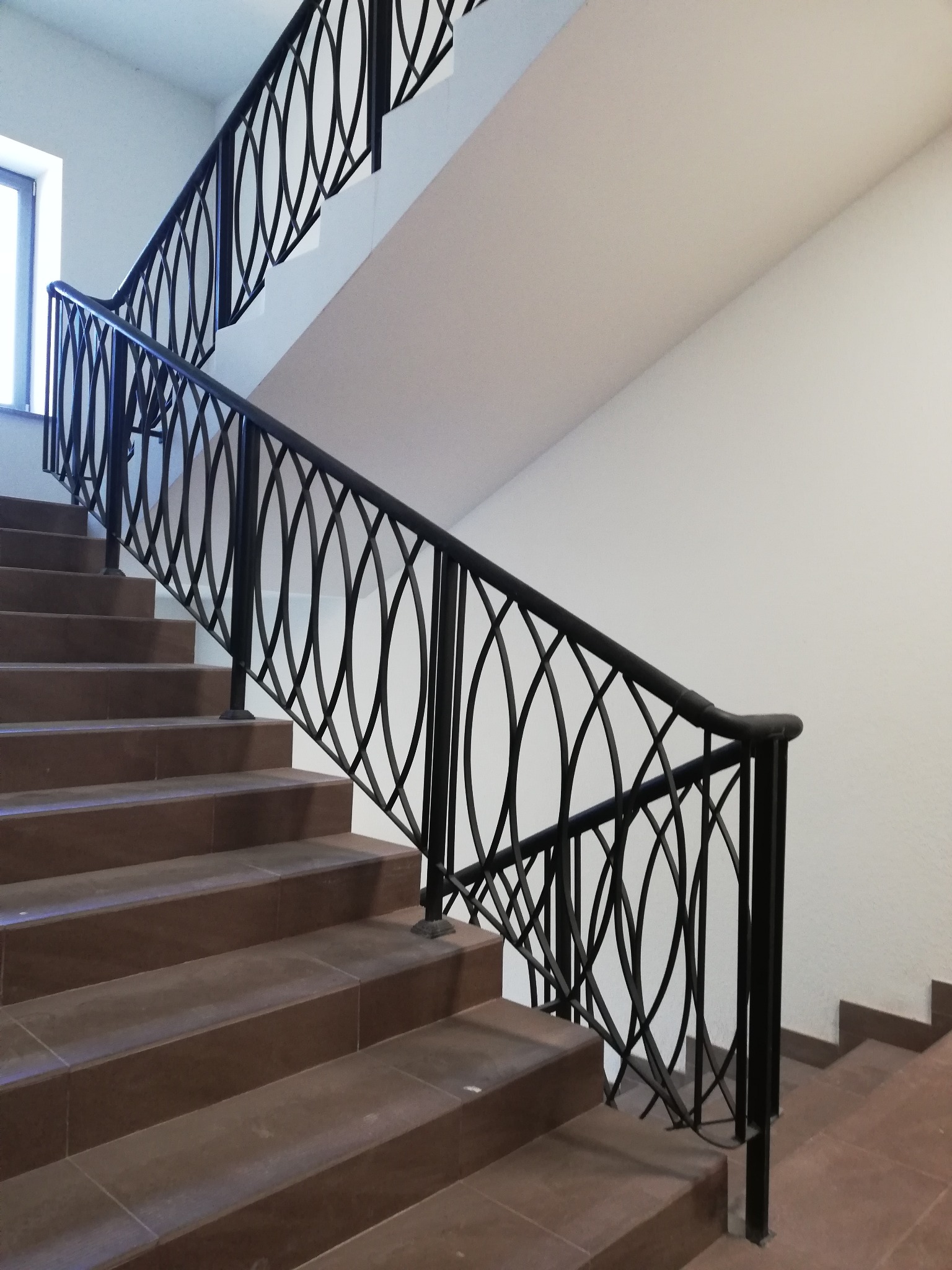 Klatka schodowa z brązowymi stopniami i czarną, ozdobną balustradą z motywem eliptycznych oczek. W tle białe ściany i okno.
