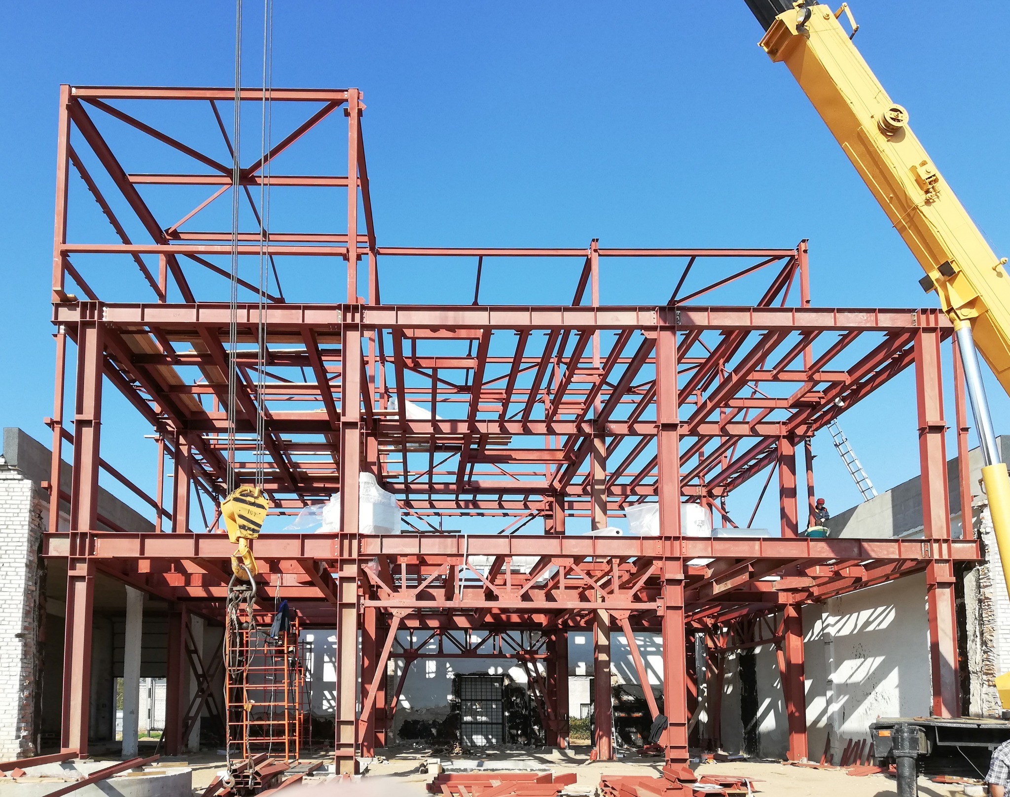 Wznoszenie szkieletu budynku z belek stalowych w kolorze czerwonym, widoczny żuraw budowlany i pracownicy na rusztowaniu, błękitne niebo w tle.