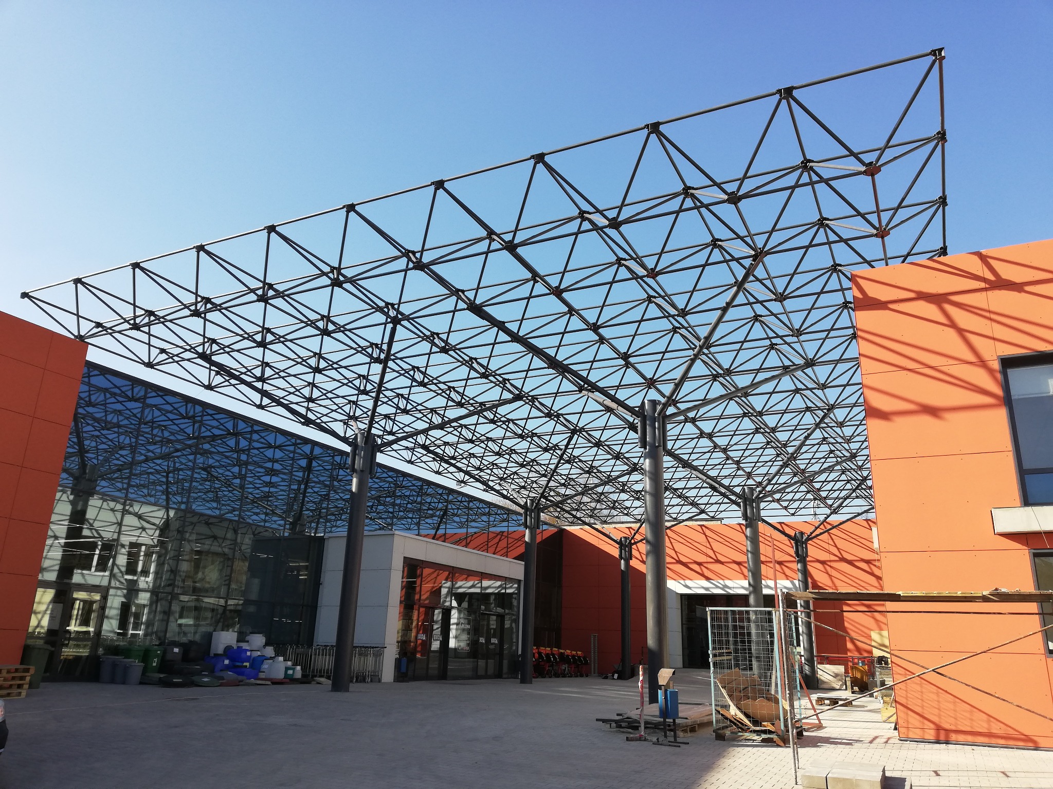 Szkielet z metalowych prętów tworzący zadaszenie nad wejściem do budynku z pomarańczową elewacją i przeszkloną fasadą, widoczne materiały budowlane i rusztowania.