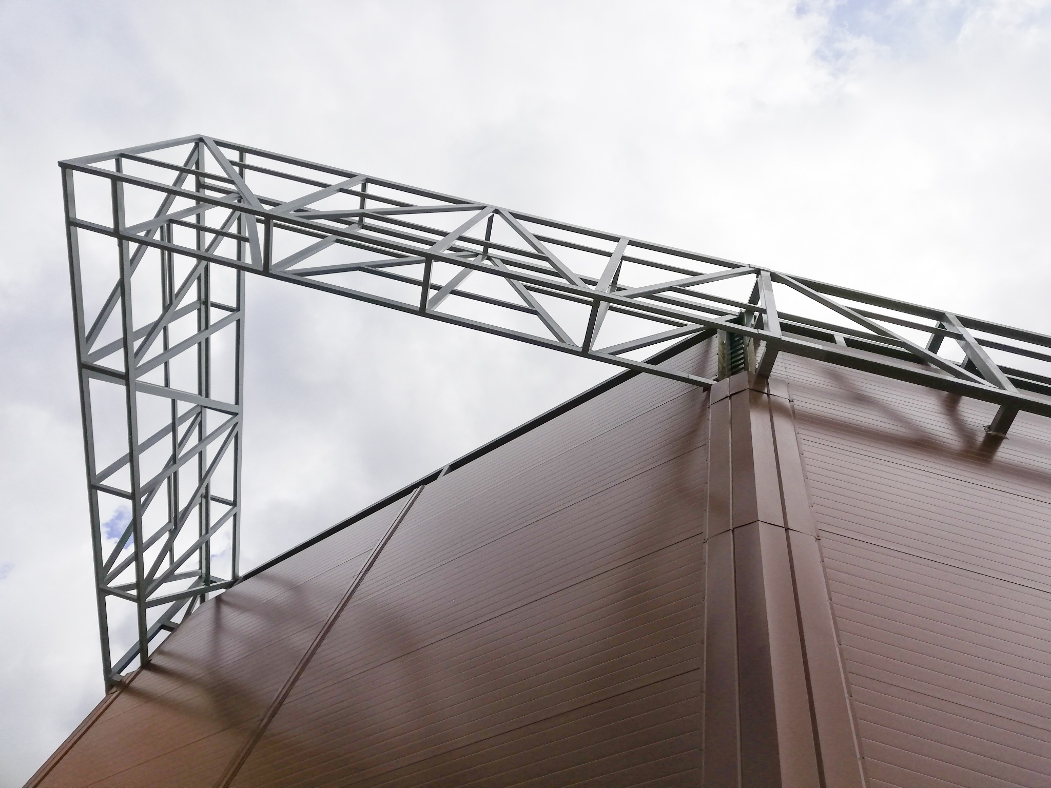 Szkielet stalowej konstrukcji budynku przemysłowego z brązową elewacją panelową, widziany z dołu na tle pochmurnego nieba.