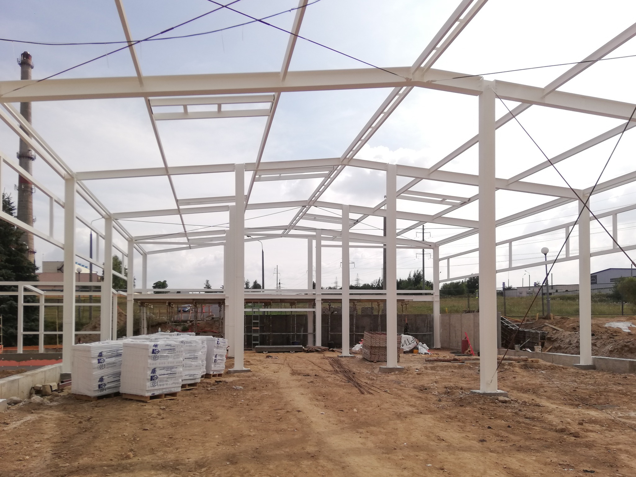 Wnętrze powstającej hali z białej stali, widoczne przygotowane materiały budowlane w foliowych opakowaniach, ziemiste podłoże i fragmenty konstrukcji w tle.