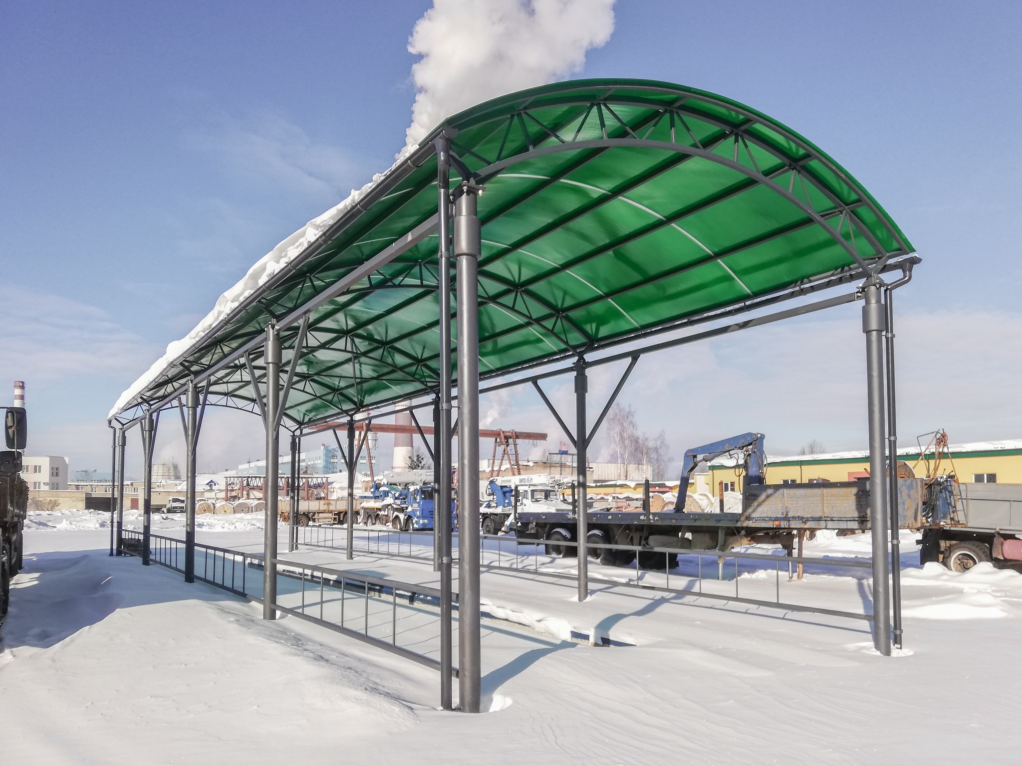 Zadaszenie z zielonego poliwęglanu na metalowej konstrukcji, pokryte śniegiem, na tle zimowego krajobrazu z zaparkowanymi ciężarówkami i kominem przemysłowym.