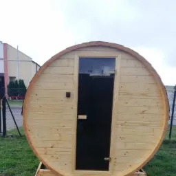 Drewniana sauna beczka z ciemnymi drzwiami na trawie, widok z przodu na tle pochmurnego nieba i budynku.