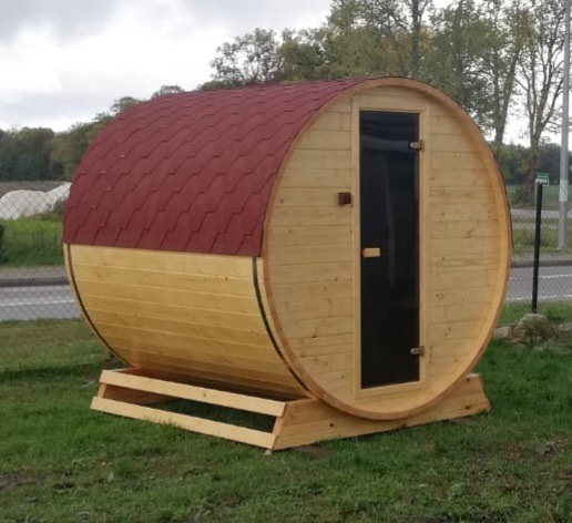 Drewniana beczka z czerwoną dachówką, prawdopodobnie gotowa do użytku, umieszczona na trawniku przed budynkiem.