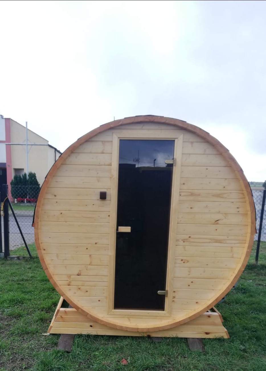 Drewniana sauna beczka z ciemnymi drzwiami na trawie, widok z przodu na tle pochmurnego nieba i budynku.